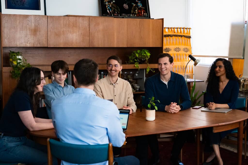 A group of six diverse professionals gathered around a wooden table in a stylish office, engaged in a collaborative meeting.