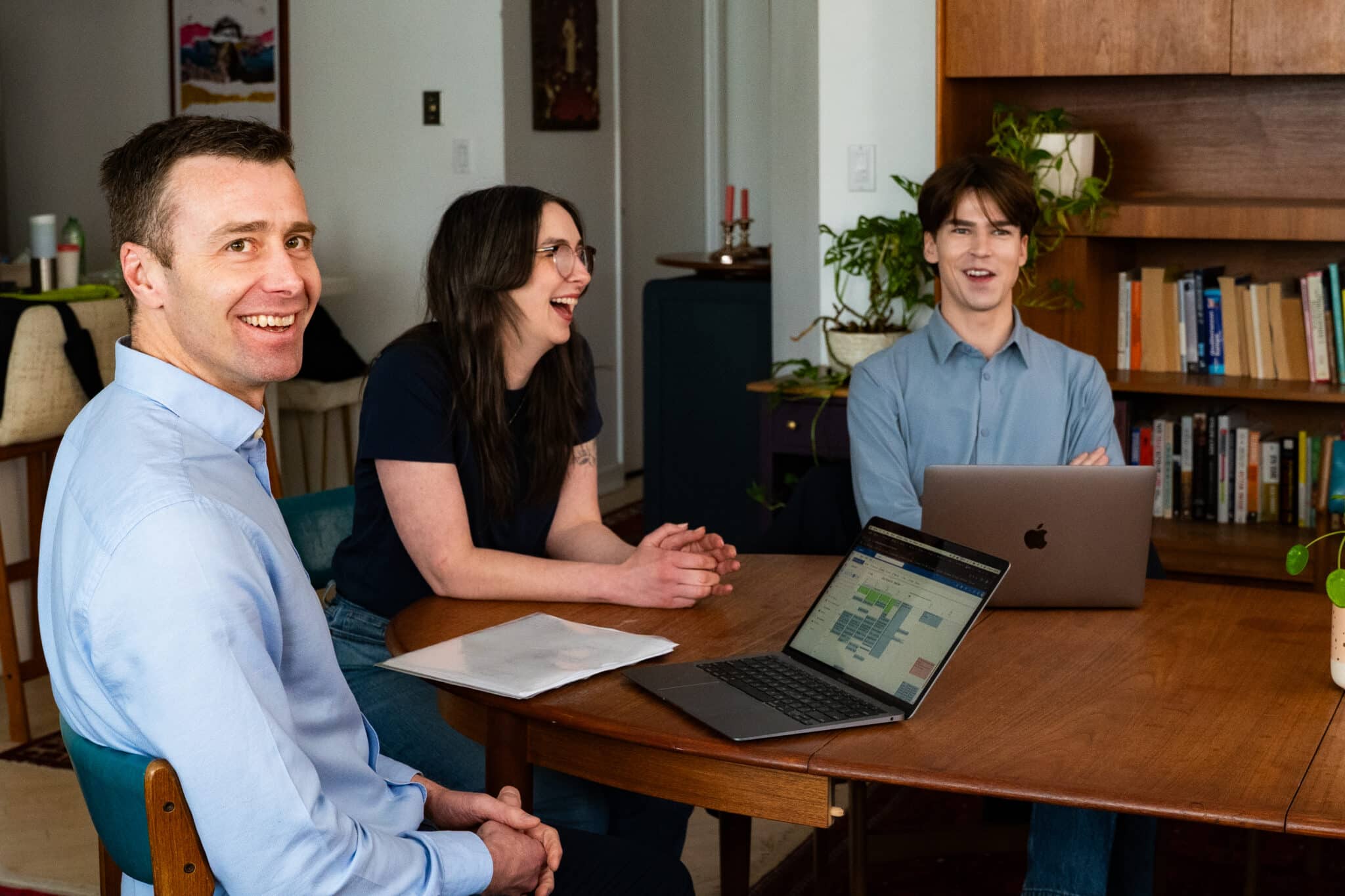 Three colleagues seated at a wooden table during a team meeting, smiling and laughing in a relaxed, collaborative atmosphere.