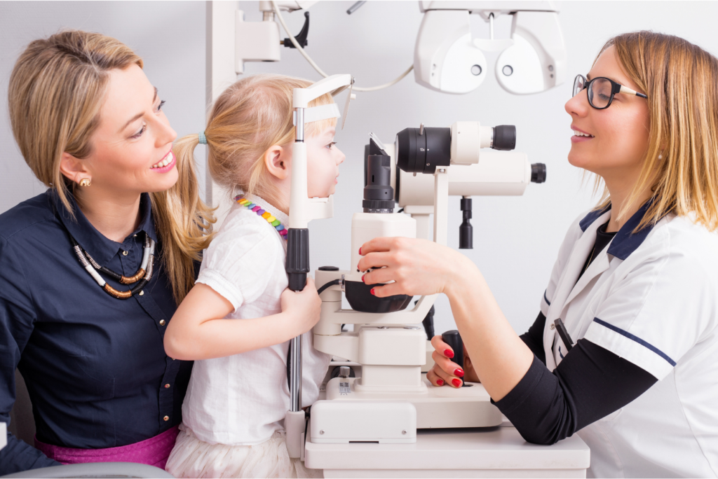 Female optometrist wearing glasses conducting a slit lamp eye examination on a young girl while her smiling mother looks on in an optometry clinic