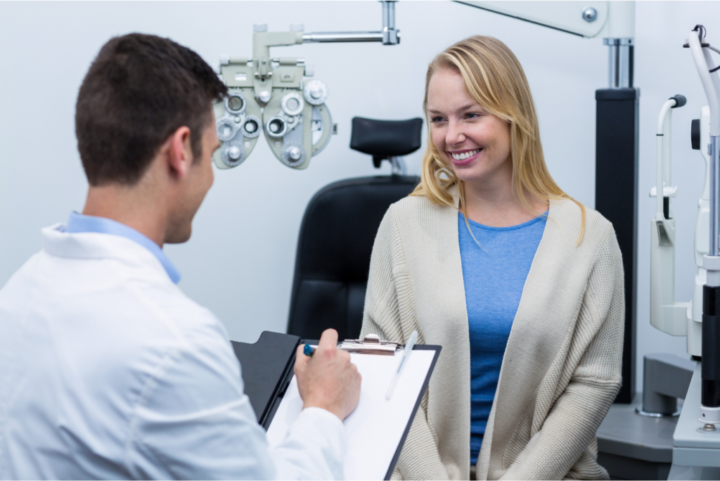 Male optometrist in a white lab coat writing notes on a clipboard while consulting with a smiling female patient in an eye exam room with a phoropter in the background