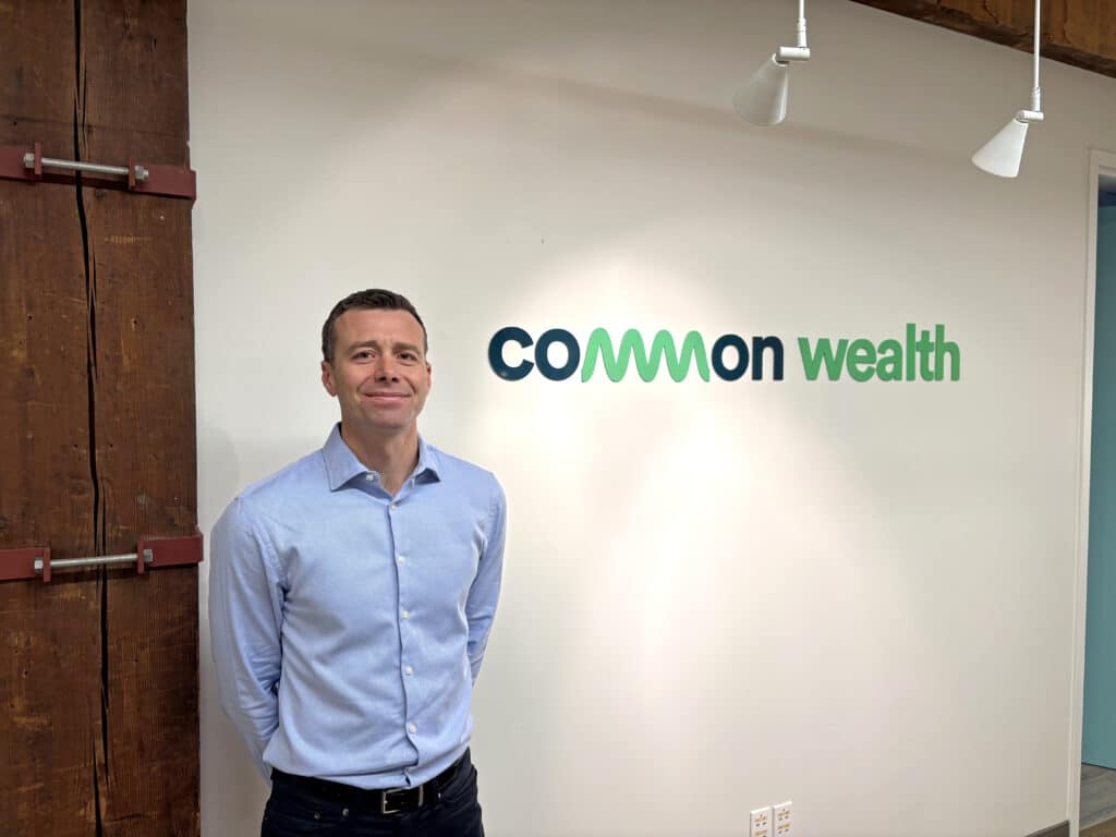 Alex Mazer, co-founder and CEO of Common Wealth, standing in front of the Common Wealth logo at the company's Toronto office.