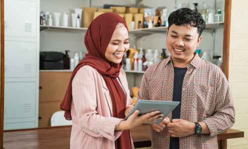 Two Muslim entrepreneurs discussing investment strategies while using a tablet in a business setting. The woman, wearing a maroon hijab and a pink blazer, smiles as she shows information on the tablet to the man in a checkered shirt.