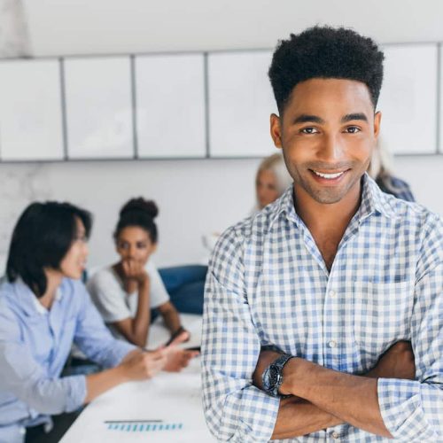 Glad young man with african hairstyle posing with arms crossed in his office with other employees on background. Male manager in blue shirt smiling during conference at workplace.. Group retirement savings plan